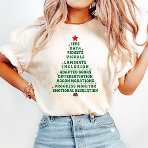 May include: Cream-colored t-shirt with a green Christmas tree design. The tree is made of words like "IEPS," "Data," and "Inclusion," topped with a red star. The shirt is paired with blue jeans.