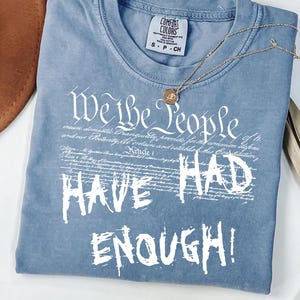 Puede incluir: Una camiseta azul Comfort Colors con el texto "We the People HAVE HAD ENOUGH!" en blanco. Un collar dorado con un colgante descansa sobre la camiseta. Un sombrero marr&oacute;n y hojas de palma secas est&aacute;n en el fondo.
