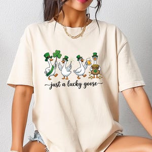 Op de afbeelding: Beige T-shirt met een afbeelding van vier cartoon ganzen gekleed in St. Patrick's Day kleding, inclusief groene hoeden en klavers. De tekst "just a lucky goose" staat onder de ganzen in een cursief lettertype.