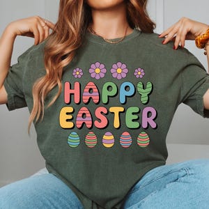 May include: Dark green t-shirt with the words "HAPPY EASTER" in colorful, bubble letters. The design includes flowers and Easter eggs. The shirt is made of a soft material and is suitable for casual wear.