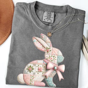Può includere: Una maglietta grigia con un design a coniglio patchwork con motivi floreali rosa, blu e bianchi. Il coniglio ha un fiocco rosa con una gemma turchese. Una collana d'argento con un ciondolo è sulla maglietta.