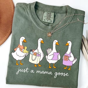 May include: Olive green t-shirt with a graphic of four cartoon geese, each with a different accessory. The text "just a mama goose" is printed below the geese. A necklace and a hat are visible in the background.