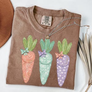 May include: A brown Comfort Colors t-shirt featuring three colorful carrot illustrations with green tops and bows. The carrots are orange, mint green, and purple with decorative patterns. A silver necklace with a pendant is on the shirt.