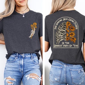 May include: Dark gray t-shirt with a graphic of a skeleton hand holding yellow flowers on the front. The back of the shirt features a design with a rib cage, flowers, and the text "Find Someone Who Grows Flowers In The Darkest Parts Of You."