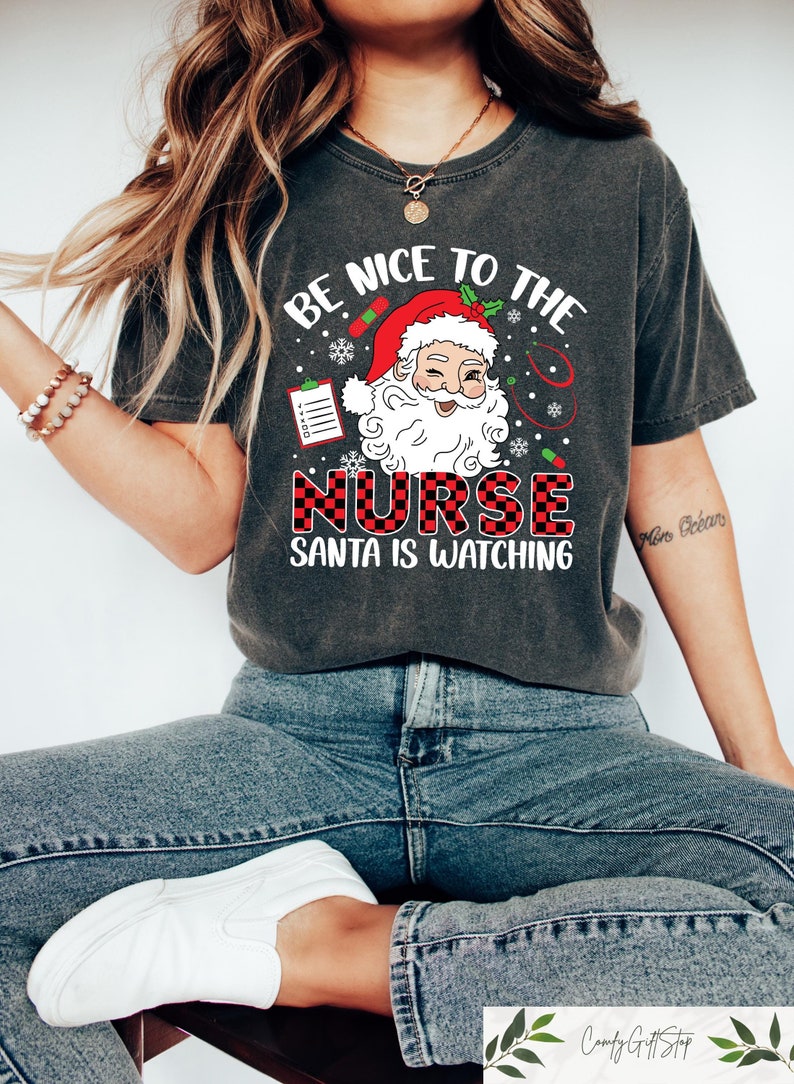 May include: A gray t-shirt with a graphic of Santa Claus wearing a red hat and a white beard. The text on the shirt reads "Be nice to the nurse Santa is watching".