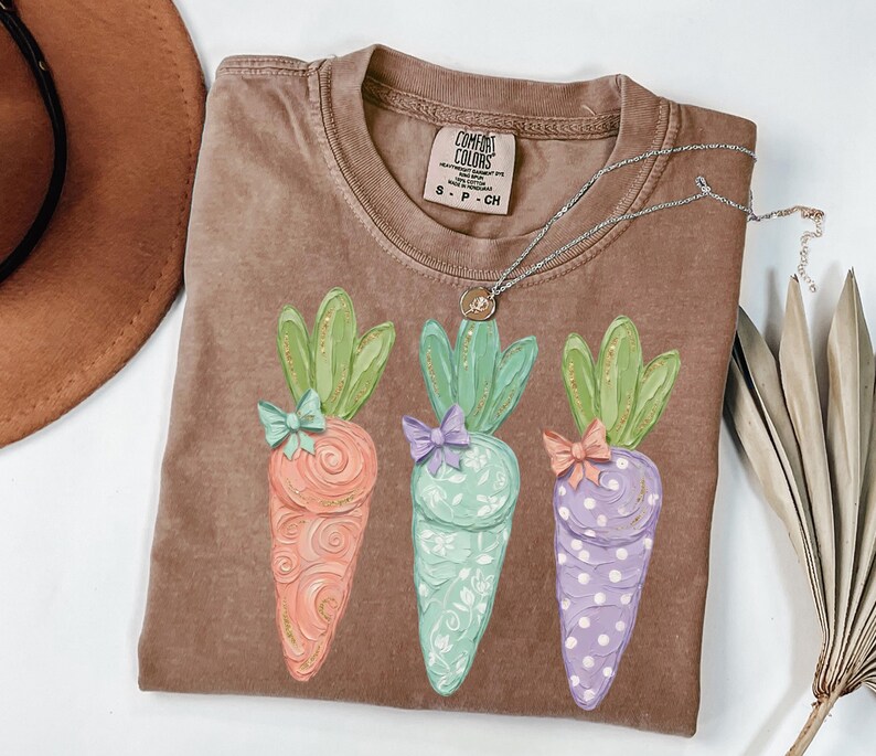 May include: A brown Comfort Colors t-shirt featuring three colorful carrot illustrations with green tops and bows. The carrots are orange, mint green, and purple with decorative patterns. A silver necklace with a pendant is on the shirt.
