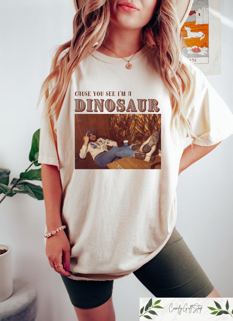May include: A white t-shirt with the text "Cause you see I'm a Dinosaur" in brown lettering. The shirt features a vintage photo of a man sitting in a chair with his feet up.