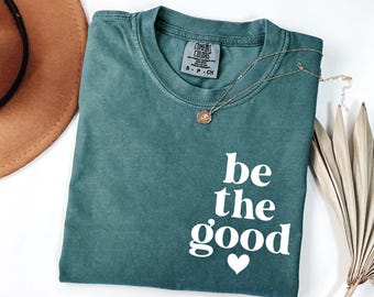 May include: A teal green short-sleeve t-shirt with a crew neckline and a Comfort Colors brand label. The shirt features white text on the lower right side that reads be the good with a small heart icon below the text. A gold necklace rests on top.