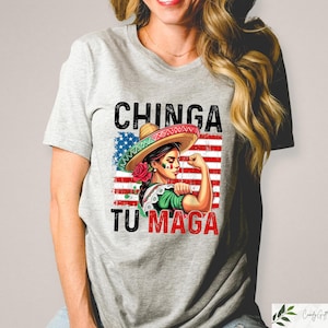May include: A light gray t-shirt featuring a graphic of a woman wearing a sombrero and flexing her arm. The design incorporates the American flag and the text "CHINGA TU MAGA" in black and red.