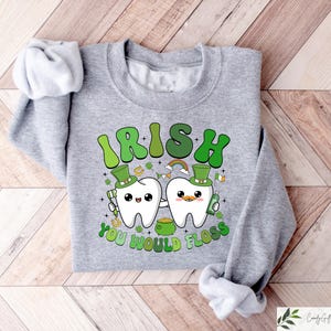 Może przedstawiać: Szara bluza z zielonym wzorem koniczyny, na kt&oacute;rej widnieje napis "Irish You Would Floss" z dwoma kresk&oacute;wkowymi zębami trzymającymi szczoteczkę do zęb&oacute;w i garnek złota.