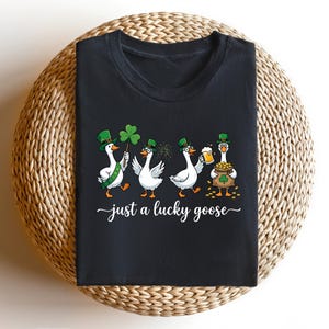 Op de afbeelding: Zwart T-shirt met een St. Patrick's Day-ontwerp. Vier cartoon ganzen zijn afgebeeld, elk met een groene hoed en met een klaver, bier of een pot goud. De tekst "just a lucky goose" is in witte sierletters geschreven.