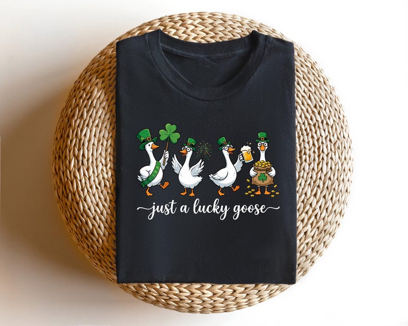 Op de afbeelding: Zwart T-shirt met een St. Patrick's Day-ontwerp. Vier cartoon ganzen zijn afgebeeld, elk met een groene hoed en met een klaver, bier of een pot goud. De tekst "just a lucky goose" is in witte sierletters geschreven.