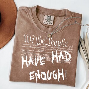 Puede incluir: Camiseta marr&oacute;n con el texto blanco "We the People HAVE HAD ENOUGH!". La camiseta tiene cuello redondo y una etiqueta peque&ntilde;a. Un collar plateado con un colgante est&aacute; drapeado sobre la camiseta.