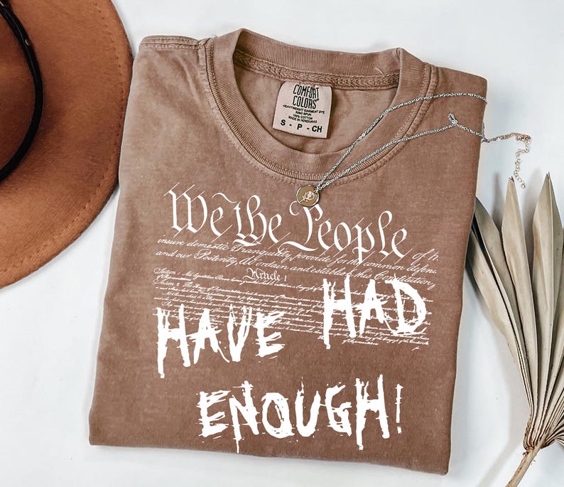 Puede incluir: Camiseta marr&oacute;n con el texto blanco "We the People HAVE HAD ENOUGH!". La camiseta tiene cuello redondo y una etiqueta peque&ntilde;a. Un collar plateado con un colgante est&aacute; drapeado sobre la camiseta.