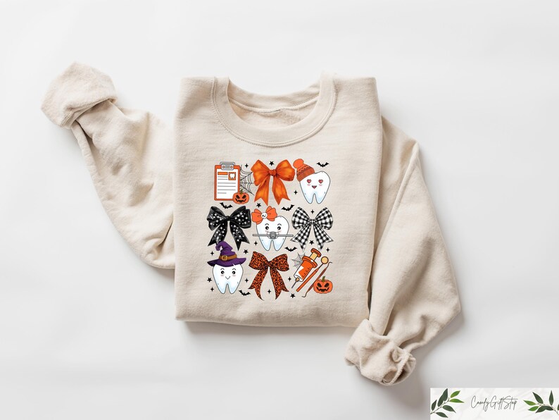 May include: A beige sweatshirt with a Halloween-themed design featuring cartoon teeth with bows, a witch's hat, a dental chart, and other dental-related imagery.