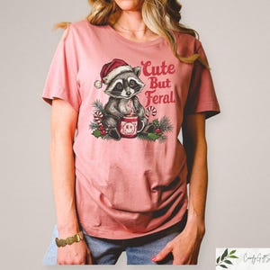 Pu&ograve; includere: Una maglietta rosa con una grafica di un procione che indossa un cappello da Babbo Natale e tiene una tazza. Il testo "Cute But Feral" &egrave; stampato sulla maglietta.