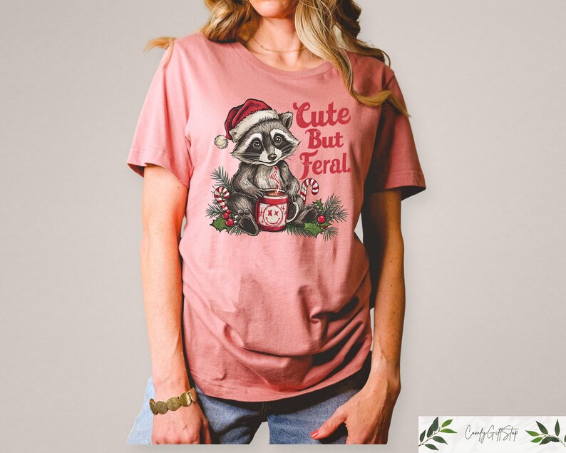 Pu&ograve; includere: Una maglietta rosa con una grafica di un procione che indossa un cappello da Babbo Natale e tiene una tazza. Il testo "Cute But Feral" &egrave; stampato sulla maglietta.