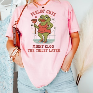以下が含まれることがあります： ピンク色のTシャツ。カエルのイラストがラバーカップを持っています。Tシャツのテキストは「Feelin' Cute Might Clog The Toilet Later」と書かれています。カエルは赤いドレスとハイヒールを履いています。ユーモラスでカジュアルなデザイン。