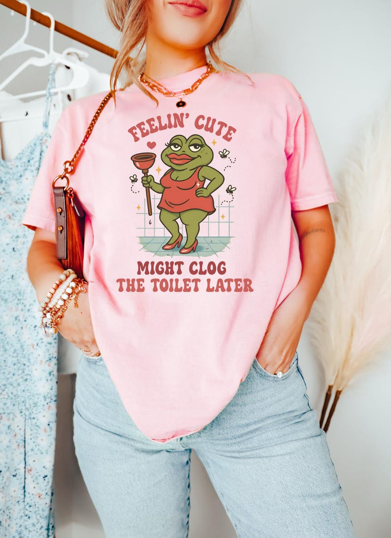 以下が含まれることがあります： ピンク色のTシャツ。カエルのイラストがラバーカップを持っています。Tシャツのテキストは「Feelin' Cute Might Clog The Toilet Later」と書かれています。カエルは赤いドレスとハイヒールを履いています。ユーモラスでカジュアルなデザイン。