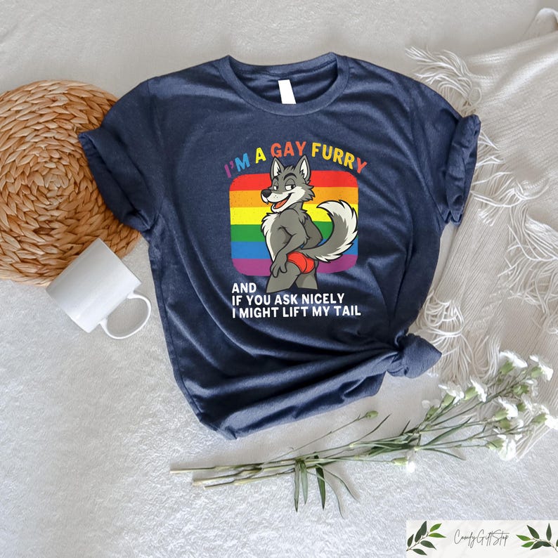 May include: Navy blue t-shirt with the text "I'M A GAY FURRY" and a cartoon wolf character. The wolf is standing in front of a rainbow-colored background. The text "AND IF YOU ASK NICELY I MIGHT LIFT MY TAIL" is below the wolf.