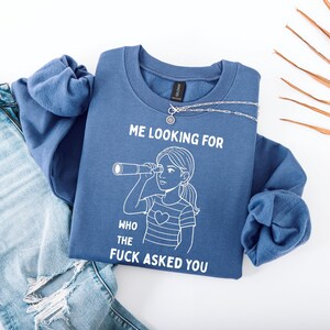 Op de afbeelding: Een blauwe sweatshirt met de tekst "ME LOOKING FOR WHO THE FUCK ASKED YOU" en een afbeelding van een persoon die door een telescoop kijkt. De sweatshirt wordt gecombineerd met een lichtblauwe jeans en een zilveren ketting.