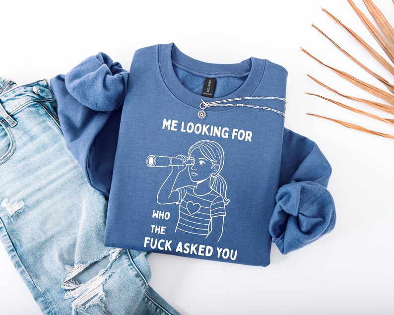 Op de afbeelding: Een blauwe sweatshirt met de tekst "ME LOOKING FOR WHO THE FUCK ASKED YOU" en een afbeelding van een persoon die door een telescoop kijkt. De sweatshirt wordt gecombineerd met een lichtblauwe jeans en een zilveren ketting.
