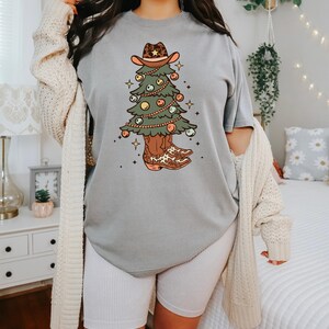 Pu&ograve; includere: Una maglietta grigia con una grafica di un albero di Natale che indossa un cappello da cowboy e stivali. L'albero &egrave; decorato con luci e ornamenti.
