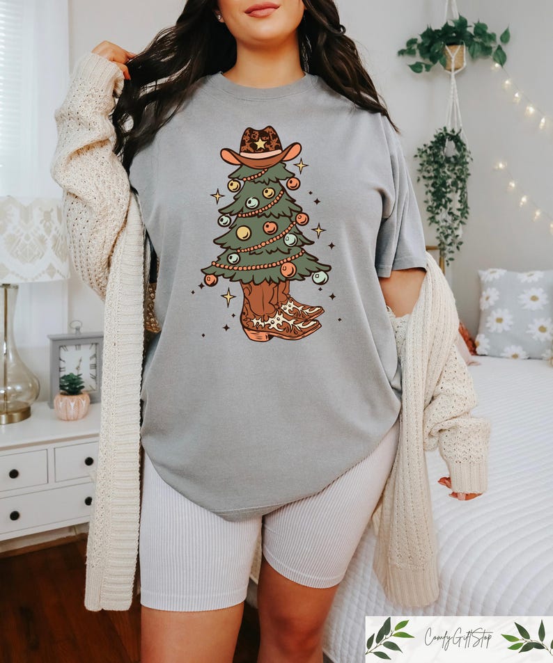 Pu&ograve; includere: Una maglietta grigia con una grafica di un albero di Natale che indossa un cappello da cowboy e stivali. L'albero &egrave; decorato con luci e ornamenti.