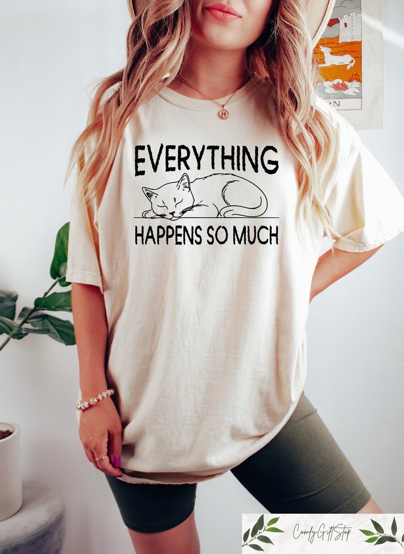K&ouml;nnte beinhalten: Cremefarbenes T-Shirt mit dem Text "EVERYTHING HAPPENS SO MUCH" und einer schwarzen Illustration einer schlafenden Katze. Das Shirt hat eine lockere Passform und kurze &Auml;rmel.