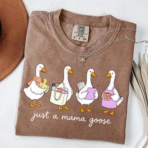 May include: Brown Comfort Colors t-shirt with the phrase "just a mama goose" and a graphic of four cartoon geese. The geese are depicted with various accessories, including a scarf, shopping bag, clipboard, and apron.