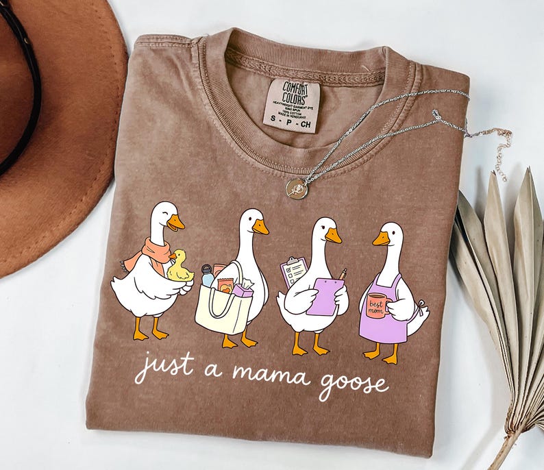 May include: Brown Comfort Colors t-shirt with the phrase "just a mama goose" and a graphic of four cartoon geese. The geese are depicted with various accessories, including a scarf, shopping bag, clipboard, and apron.
