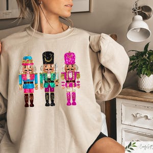 May include: A beige sweatshirt with a graphic of three nutcracker figurines. The nutcrackers are wearing blue, green, and pink outfits with gold accents. The sweatshirt has a relaxed fit and a slightly puffed sleeve.