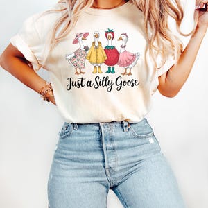 May include: A white t-shirt with a graphic of four geese dressed in different outfits. The geese are wearing a pink dress, a yellow raincoat, a strawberry costume, and a floral dress. The text "Just a Silly Goose" is printed below the geese.