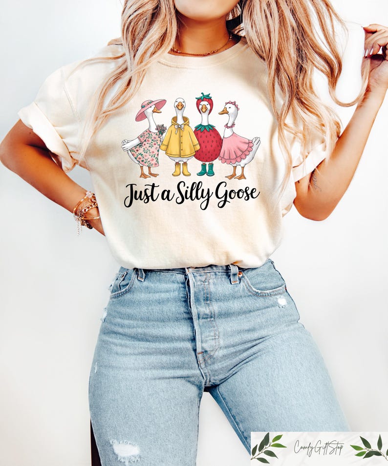 May include: A white t-shirt with a graphic of four geese dressed in different outfits. The geese are wearing a pink dress, a yellow raincoat, a strawberry costume, and a floral dress. The text "Just a Silly Goose" is printed below the geese.
