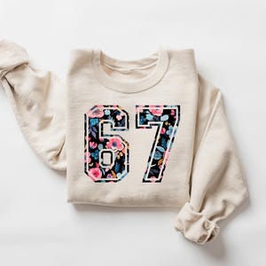 May include: A cream-colored sweatshirt with the number "67" in a floral design. The floral pattern includes pink, blue, and white flowers on a black background. The sleeves are rolled up.