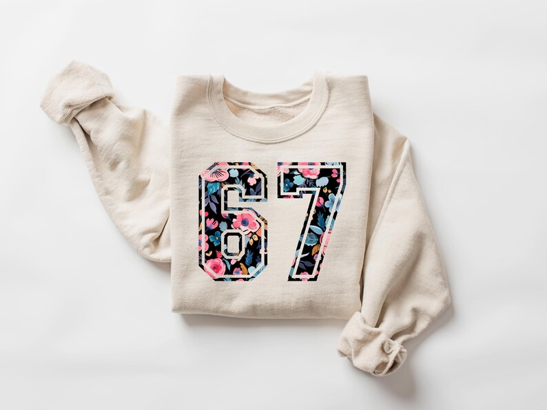 May include: A cream-colored sweatshirt with the number "67" in a floral design. The floral pattern includes pink, blue, and white flowers on a black background. The sleeves are rolled up.
