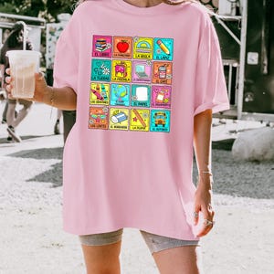 May include: A pale pink t-shirt with a grid of colourful school-related images and Spanish words. The grid includes a book, apple, scissors, backpack, and other items. The t-shirt has a loose fit.