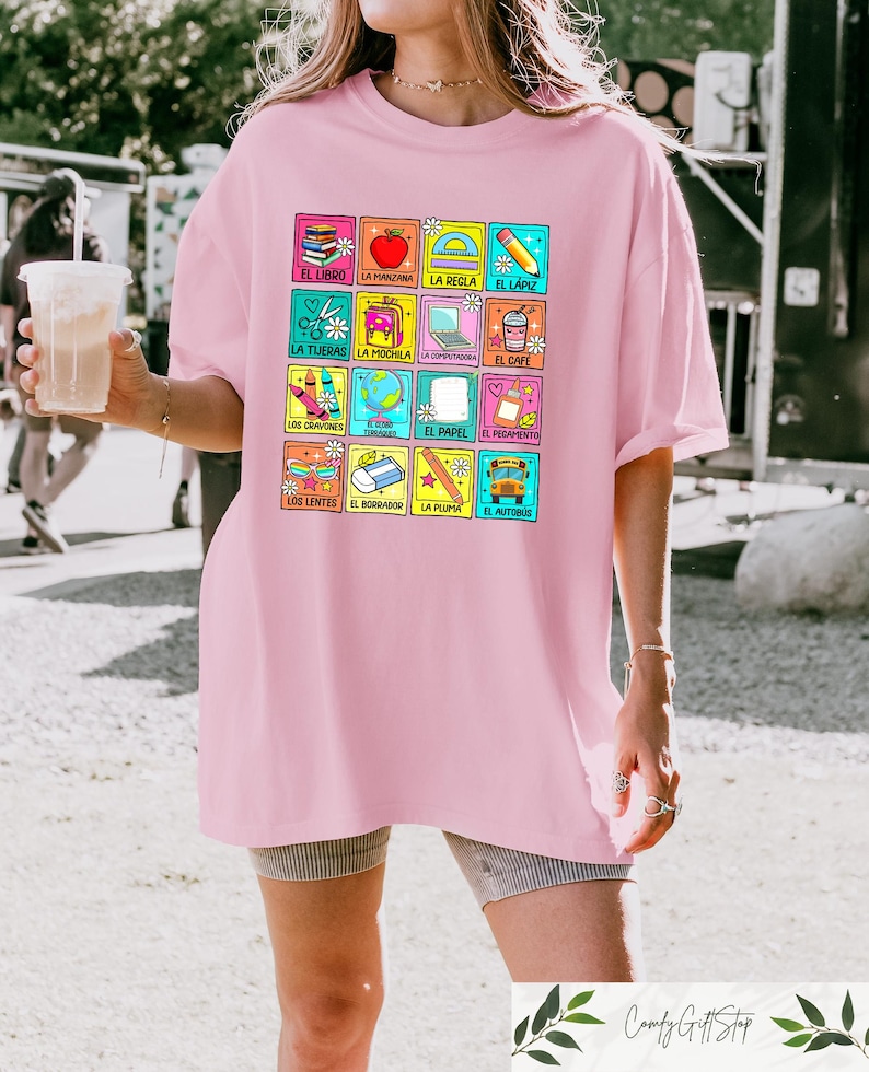 May include: A pale pink t-shirt with a grid of colourful school-related images and Spanish words. The grid includes a book, apple, scissors, backpack, and other items. The t-shirt has a loose fit.