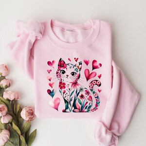 May include: A light pink sweatshirt featuring a watercolor illustration of a cat adorned with floral patterns and surrounded by pink and teal hearts. The cat's design incorporates flowers and leaves. The sweatshirt has long sleeves.