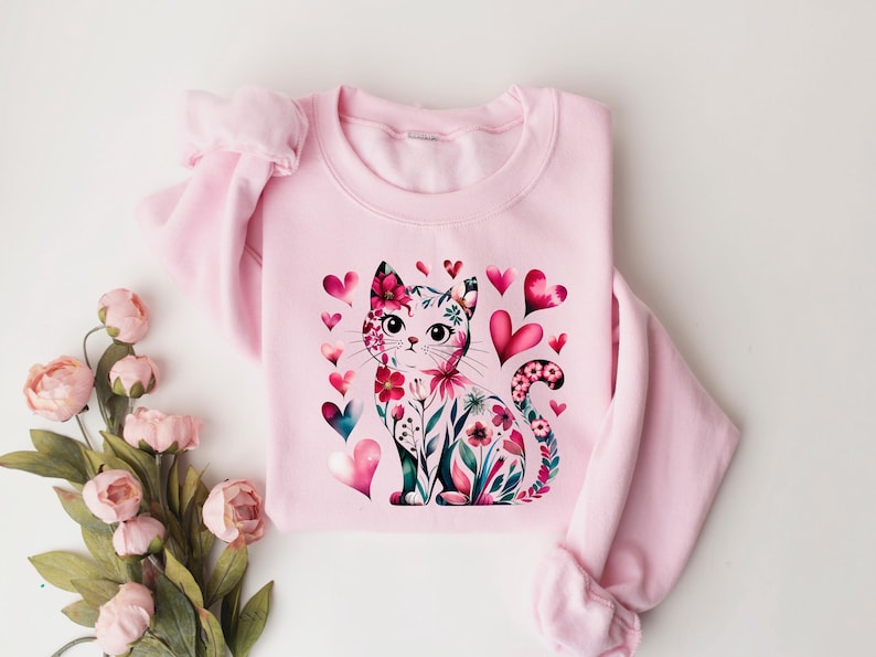 May include: A light pink sweatshirt featuring a watercolor illustration of a cat adorned with floral patterns and surrounded by pink and teal hearts. The cat's design incorporates flowers and leaves. The sweatshirt has long sleeves.
