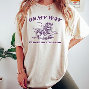 May include: A cream colored t-shirt with a purple graphic of a person riding a goose. The text "On My Way To Make The Vibe Weird" is printed on the shirt.