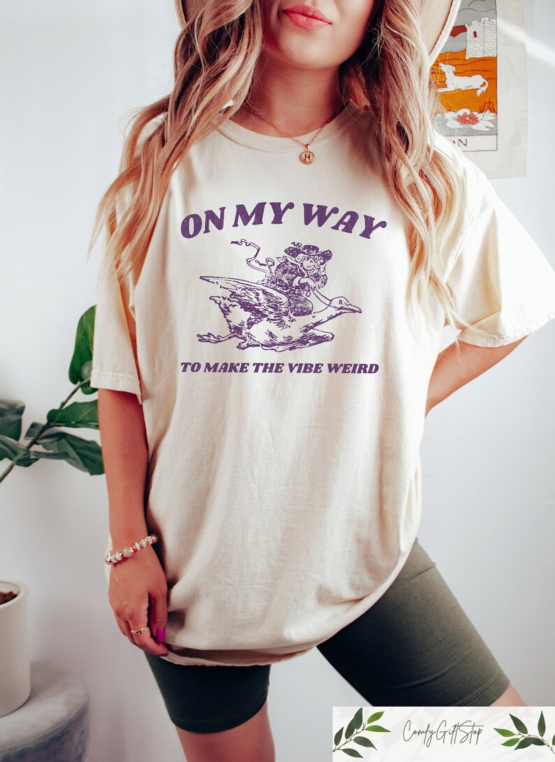 May include: A cream colored t-shirt with a purple graphic of a person riding a goose. The text "On My Way To Make The Vibe Weird" is printed on the shirt.