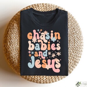 Peut inclure: T-shirt noir avec un graphisme color&eacute; qui dit "chasin babies and Jesus" avec des fleurs et des &eacute;toiles autour du texte.