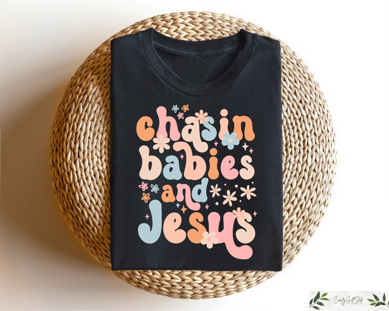 Peut inclure: T-shirt noir avec un graphisme color&eacute; qui dit "chasin babies and Jesus" avec des fleurs et des &eacute;toiles autour du texte.