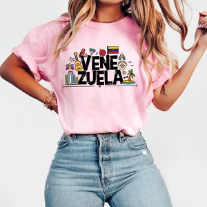 May include: A pink t-shirt with a graphic design featuring the word "VENEZUELA" in black text with colorful icons representing Venezuelan culture, including a guitar, a parrot, a flower, a flag, and a palm tree.