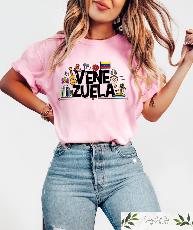 May include: A pink t-shirt with a graphic design featuring the word "VENEZUELA" in black text with colorful icons representing Venezuelan culture, including a guitar, a parrot, a flower, a flag, and a palm tree.
