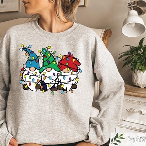 May include: A light gray sweatshirt featuring a graphic of three gnomes wearing colorful Christmas lights. The gnomes have blue, green, and red hats and white beards. The design is centered on the front of the sweatshirt.