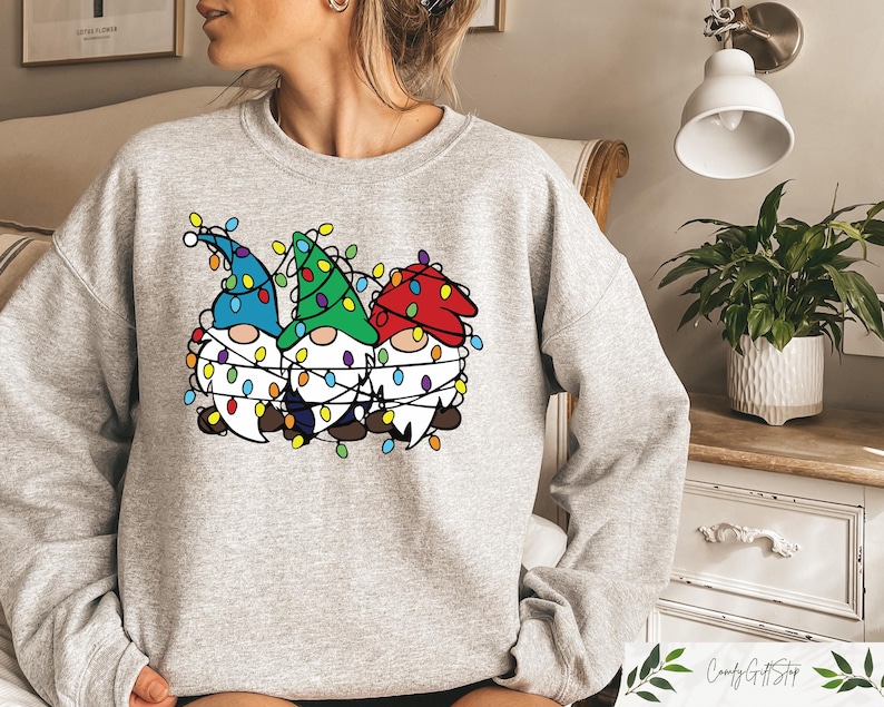 May include: A light gray sweatshirt featuring a graphic of three gnomes wearing colorful Christmas lights. The gnomes have blue, green, and red hats and white beards. The design is centered on the front of the sweatshirt.