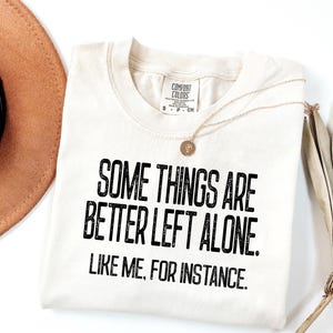 May include: Cream-colored t-shirt with the text "SOME THINGS ARE BETTER LEFT ALONE. LIKE ME, FOR INSTANCE." in black. A gold necklace with a pendant and a brown hat are also visible.