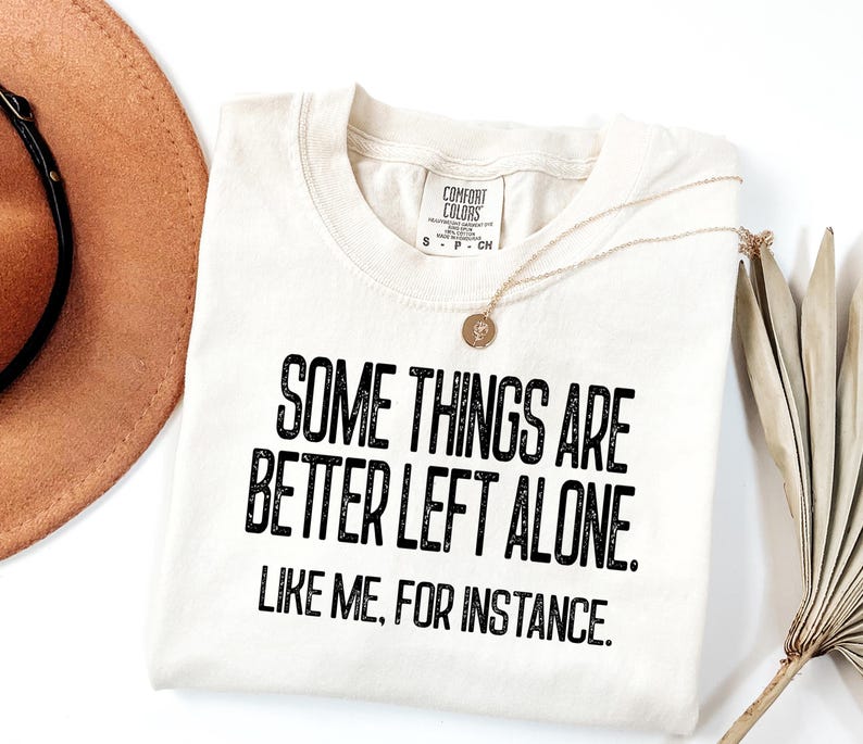 May include: Cream-colored t-shirt with the text "SOME THINGS ARE BETTER LEFT ALONE. LIKE ME, FOR INSTANCE." in black. A gold necklace with a pendant and a brown hat are also visible.